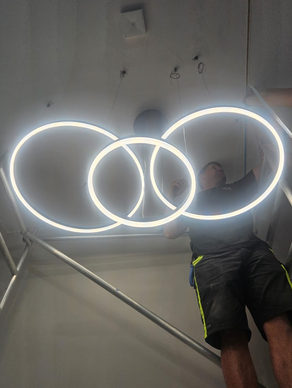 Elektryk na rusztowaniu montuje nowoczesną, okrągłą lampę sufitową LED z trzema pierścieniami. Widoczne kable i elementy mocujące.