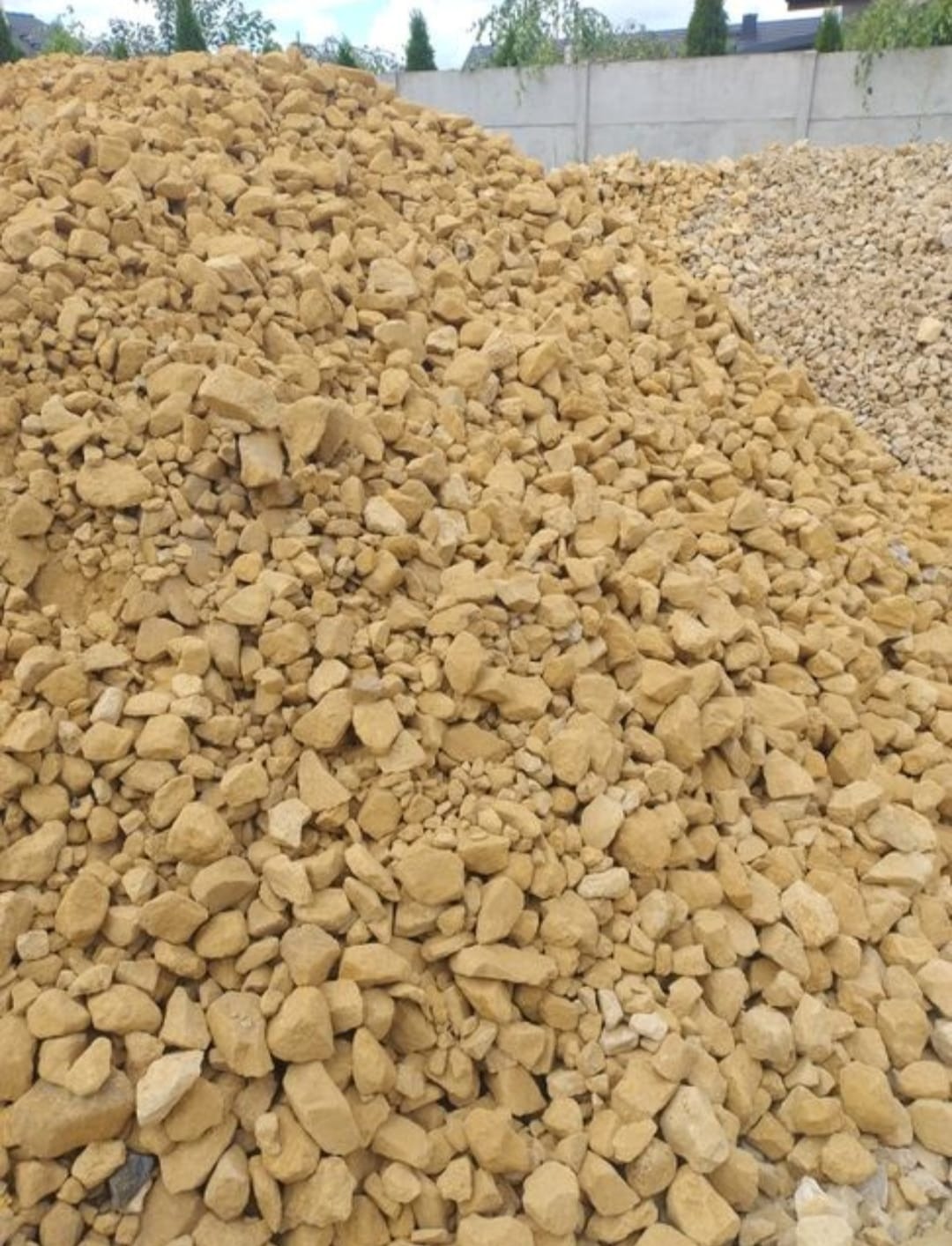 Góra piasku w ciepłym, beżowym odcieniu, z widoczną frakcją i teksturą, na tle betonowego muru i zieleni drzew, prezentowana w naturalnym oświetleniu.
