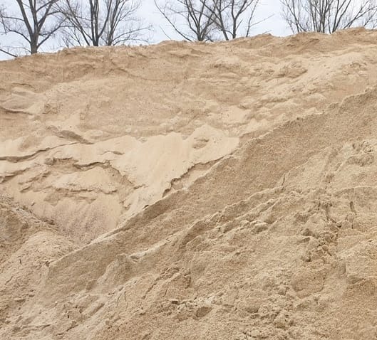 Jasny piasek budowlany w dużej pryzmie, widoczne ślady po ześlizgiwaniu się materiału. W tle drzewa bez liści.