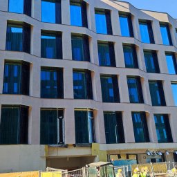 AKO GROUP - Elewacja budynku z licznymi oknami, w trakcie prac wykończeniowych. Widoczny fragment rusztowania i pracownicy w żółtych kaskach. Nowoczesna architektura.