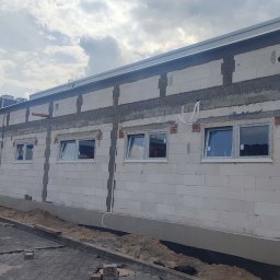 AKO GROUP - Budynek w trakcie wznoszenia z betonu komórkowego. Widoczne okna w białych ramach, niezabezpieczone przewody i mini koparka. Częściowo ułożona kostka brukowa.