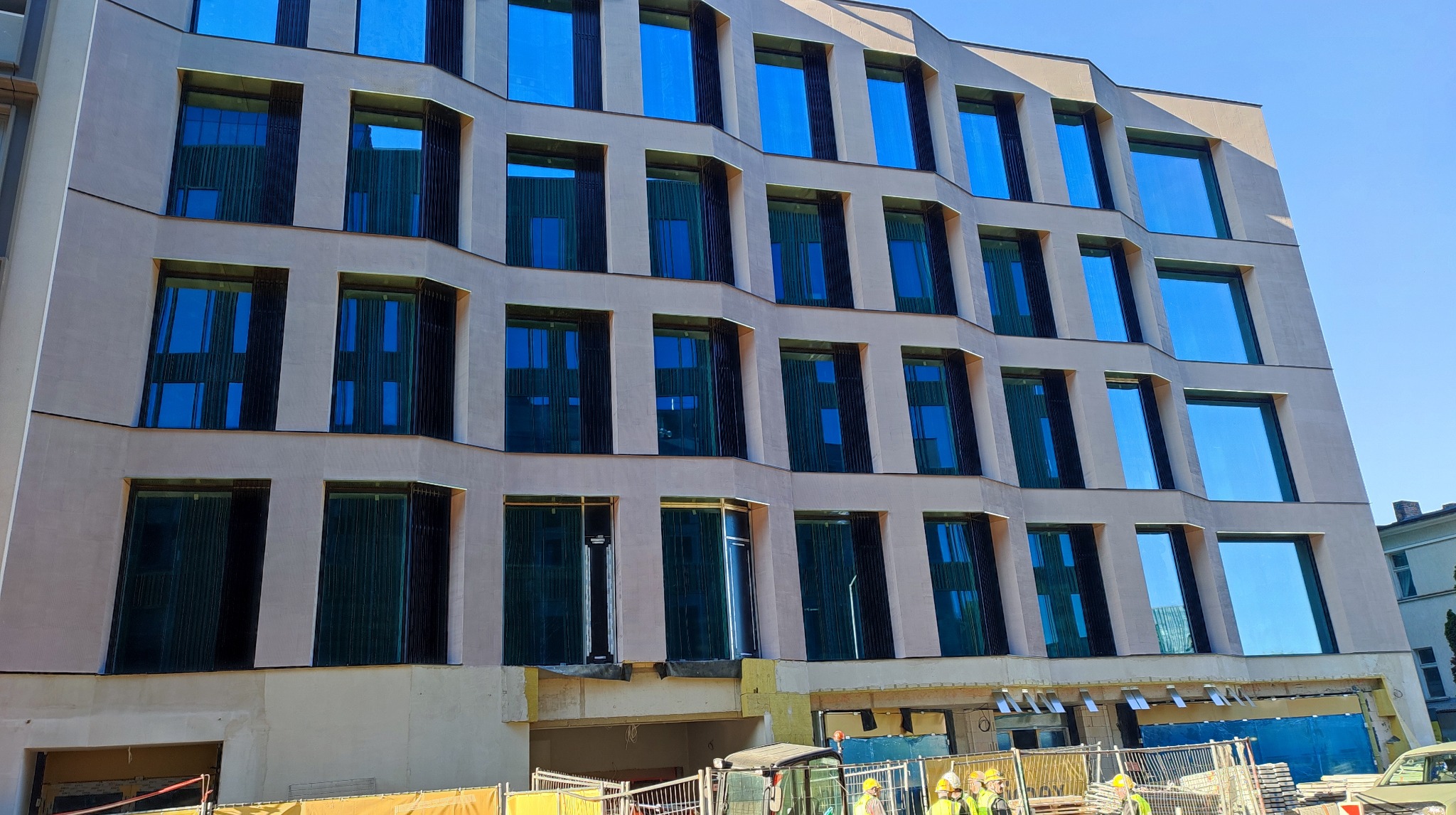 Elewacja budynku z licznymi oknami, w trakcie prac wykończeniowych. Widoczny fragment rusztowania i pracownicy w żółtych kaskach. Nowoczesna architektura.