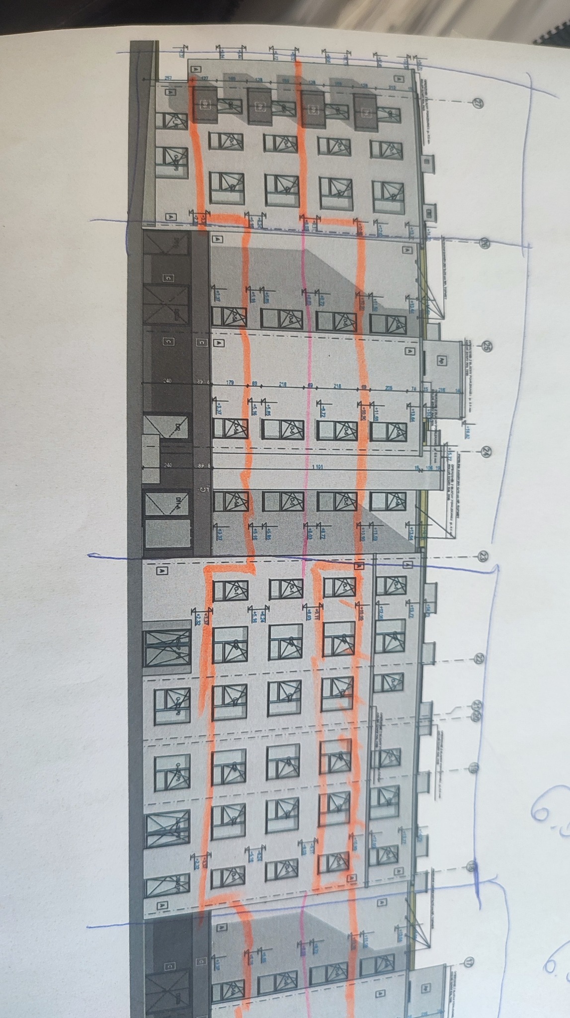 Ręcznie naniesione poprawki na planie elewacji budynku wielorodzinnego, z zaznaczonymi pionowymi liniami w kolorze pomarańczowym. Widoczne okna i wymiary.