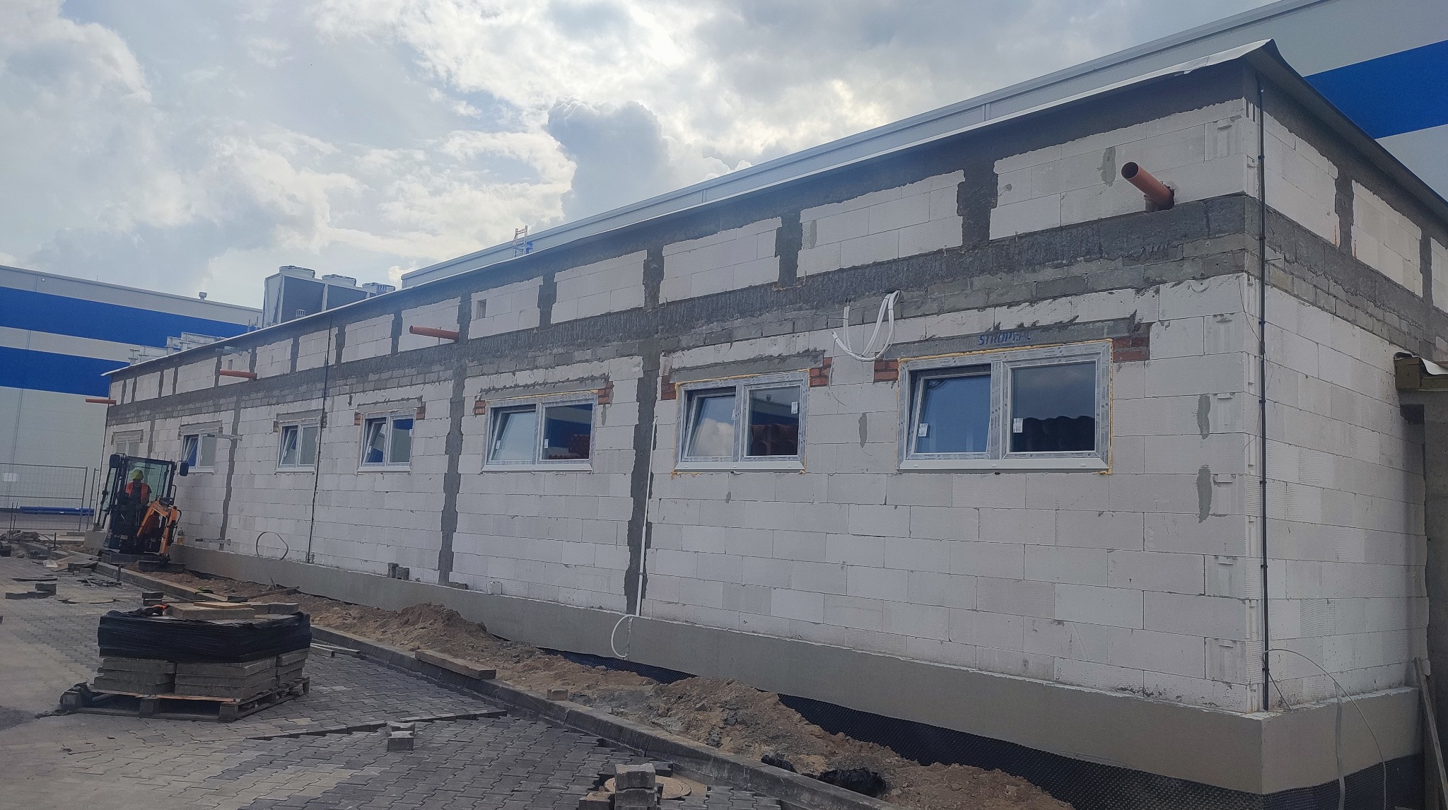 Budynek w trakcie wznoszenia z betonu komórkowego. Widoczne okna w białych ramach, niezabezpieczone przewody i mini koparka. Częściowo ułożona kostka brukowa.