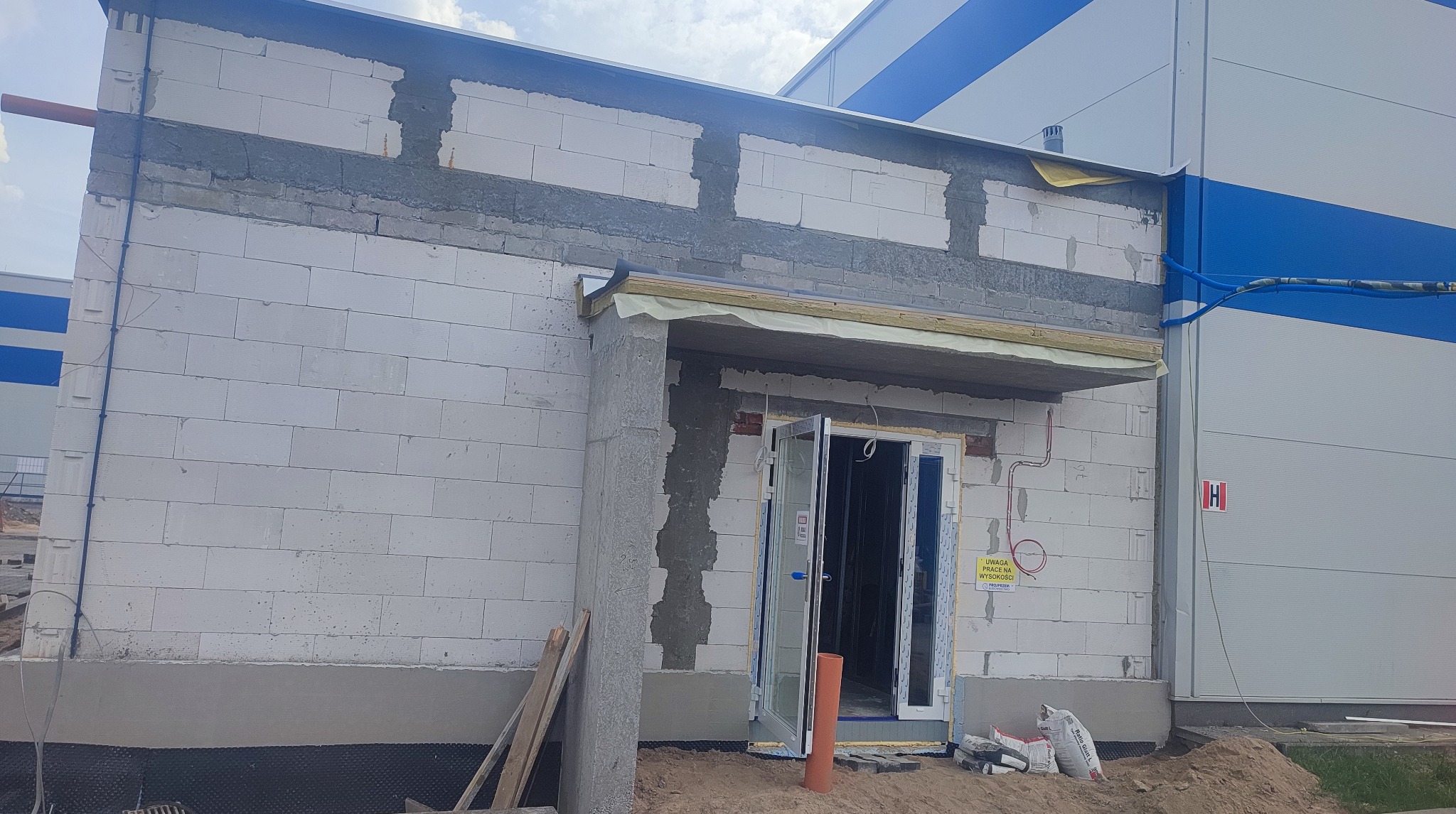 Wznoszenie ściany z białych bloczków, widoczny stan surowy z otwartymi drzwiami wejściowymi, zaprawa murarska i instalacje elektryczne. Budowa w toku.