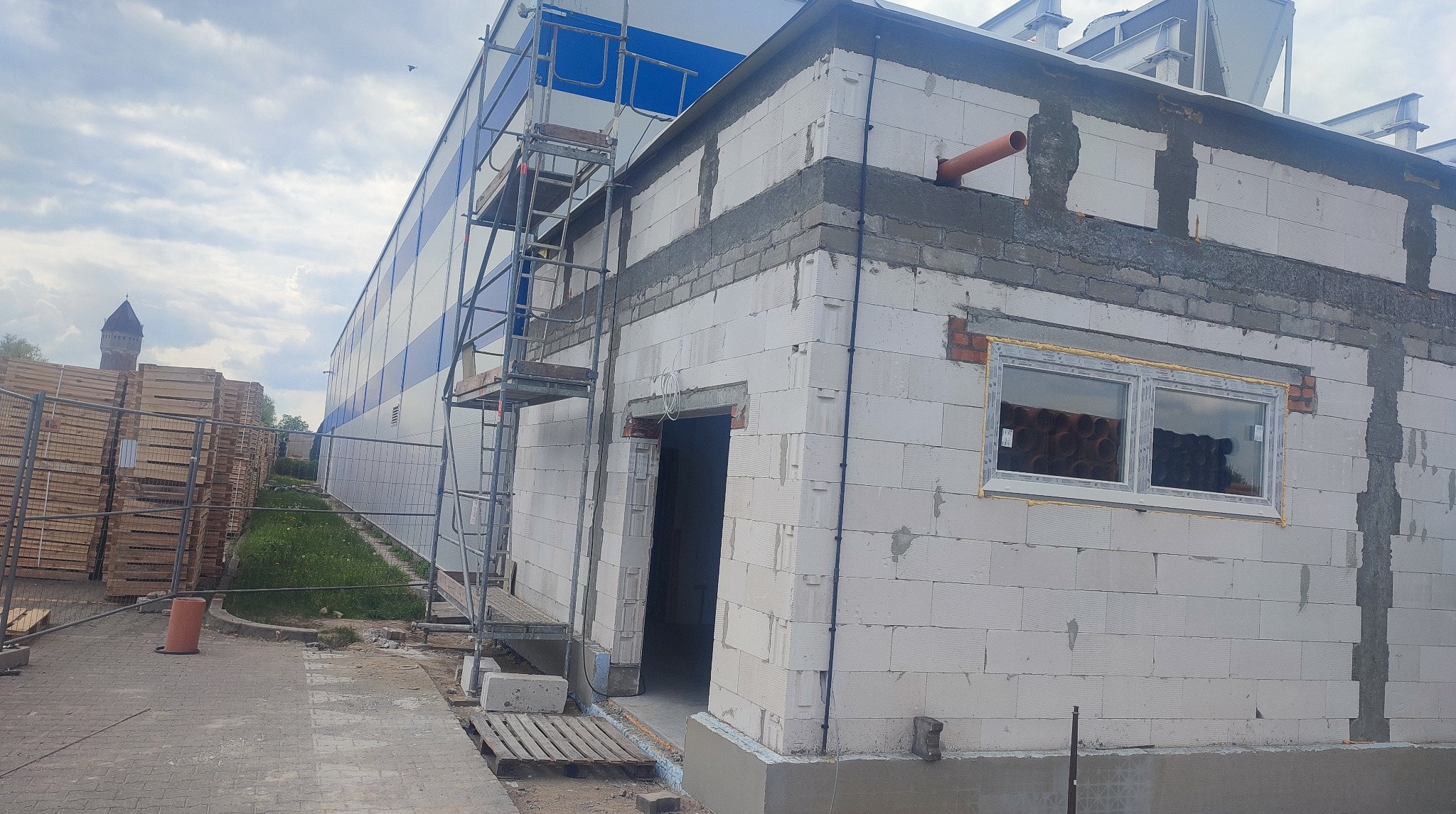 Budowa budynku z białych bloczków, widoczne rusztowanie, okno z rurami w środku, fragment elewacji hali w tle, elementy instalacji na ścianie.
