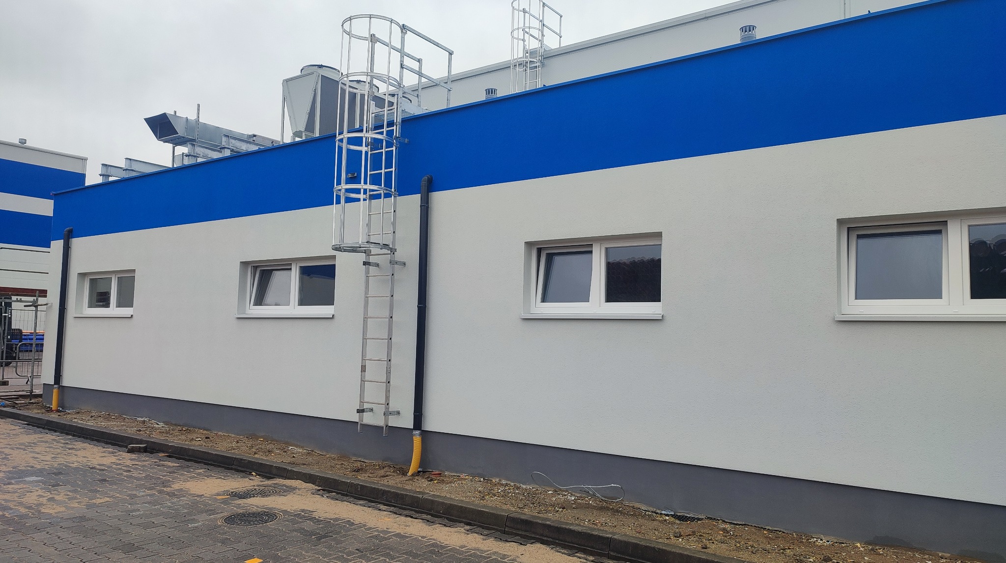Wykończona elewacja budynku z szaro-białą fasadą i niebieskim pasem na górze. Widoczne okna, drabina ewakuacyjna i instalacje wentylacyjne na dachu.