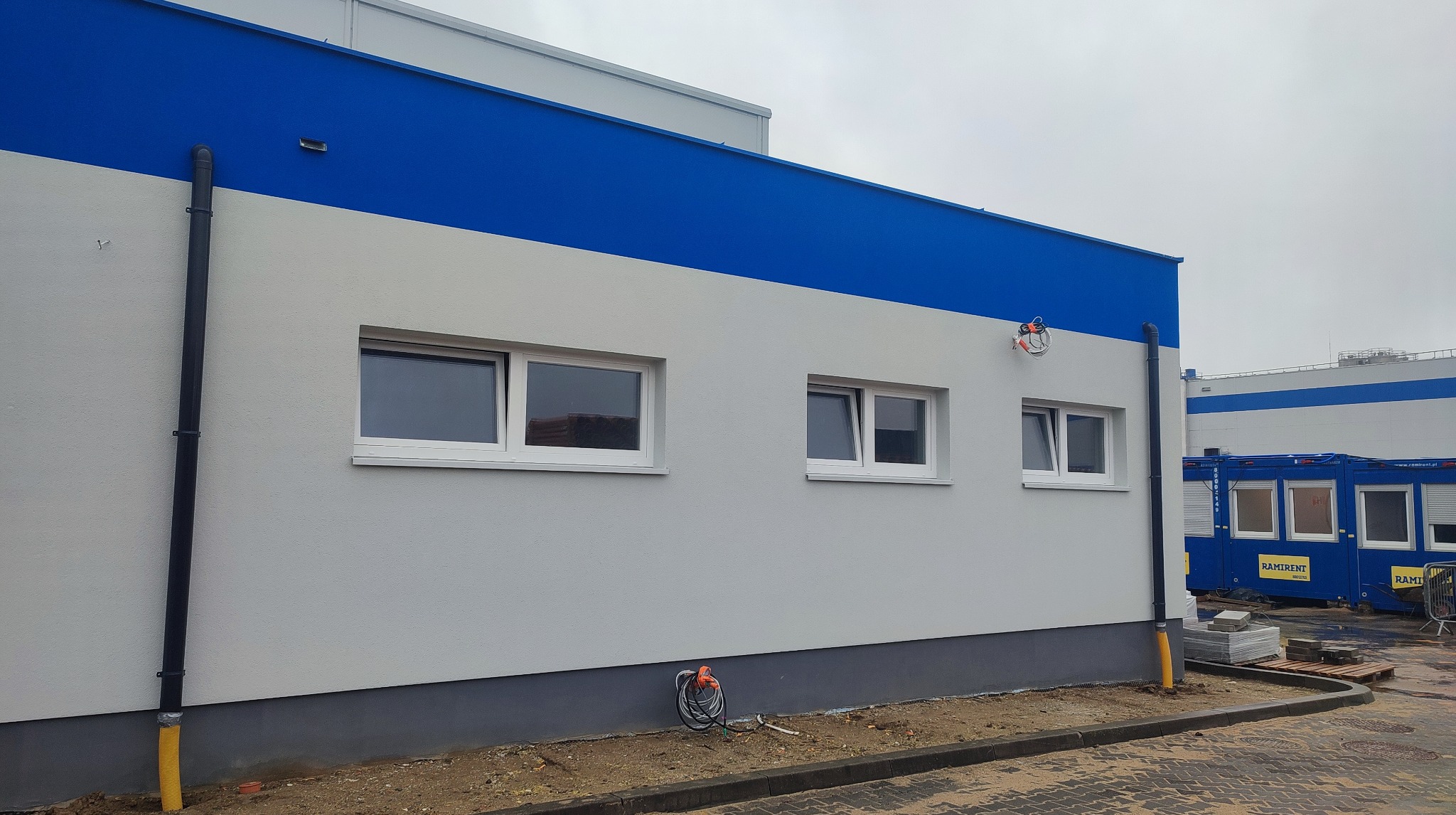 Elewacja budynku po malowaniu: szary tynk, niebieski pas na górze, białe okna. Widoczne rury spustowe i zwinięty kabel elektryczny przy ziemi.