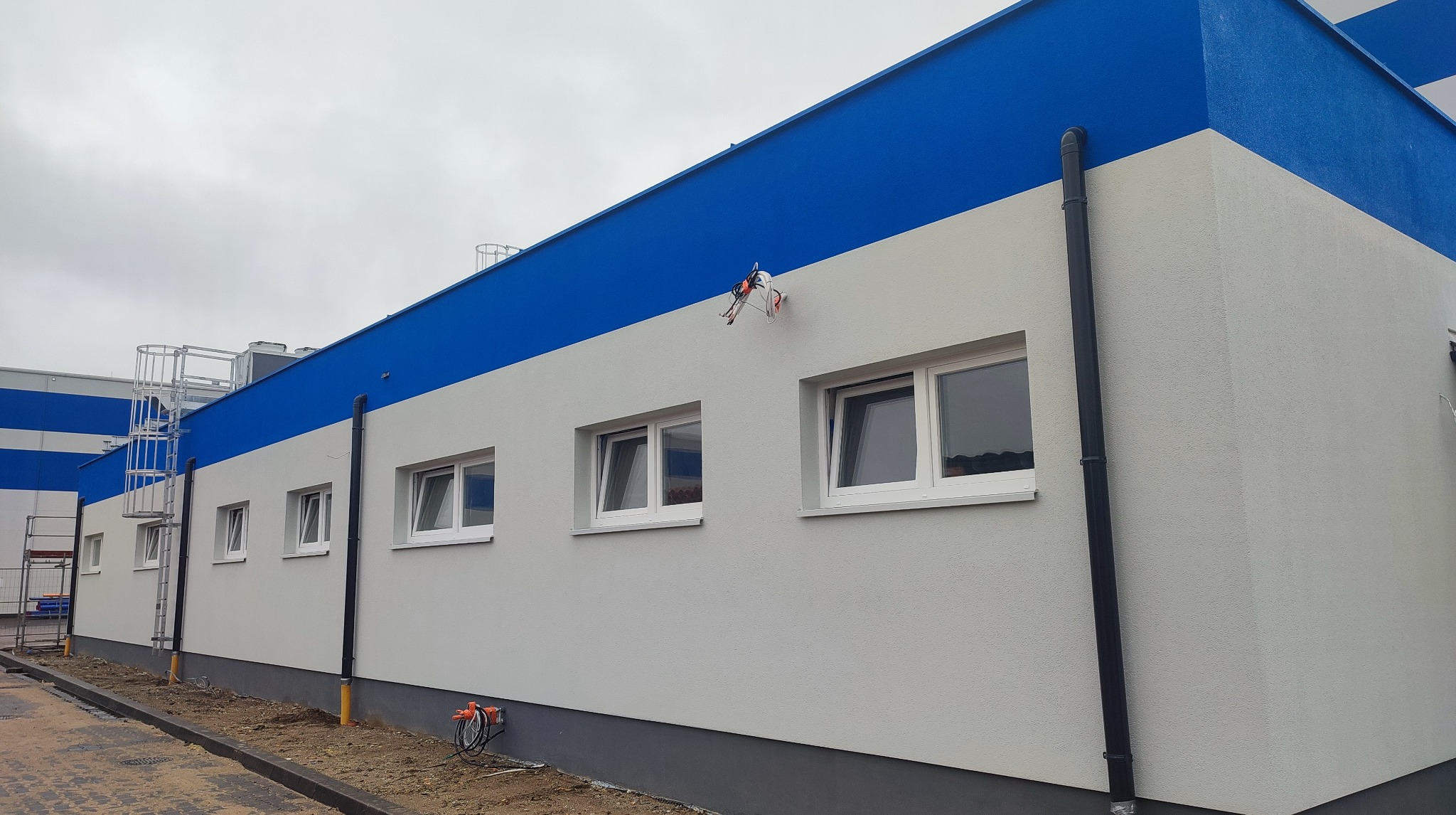 Elewacja budynku po malowaniu na biało i niebiesko. Widoczne okna, rynny, drabina i sprzęt malarski. Świeżo pomalowana fasada z wyraźną strukturą tynku.
