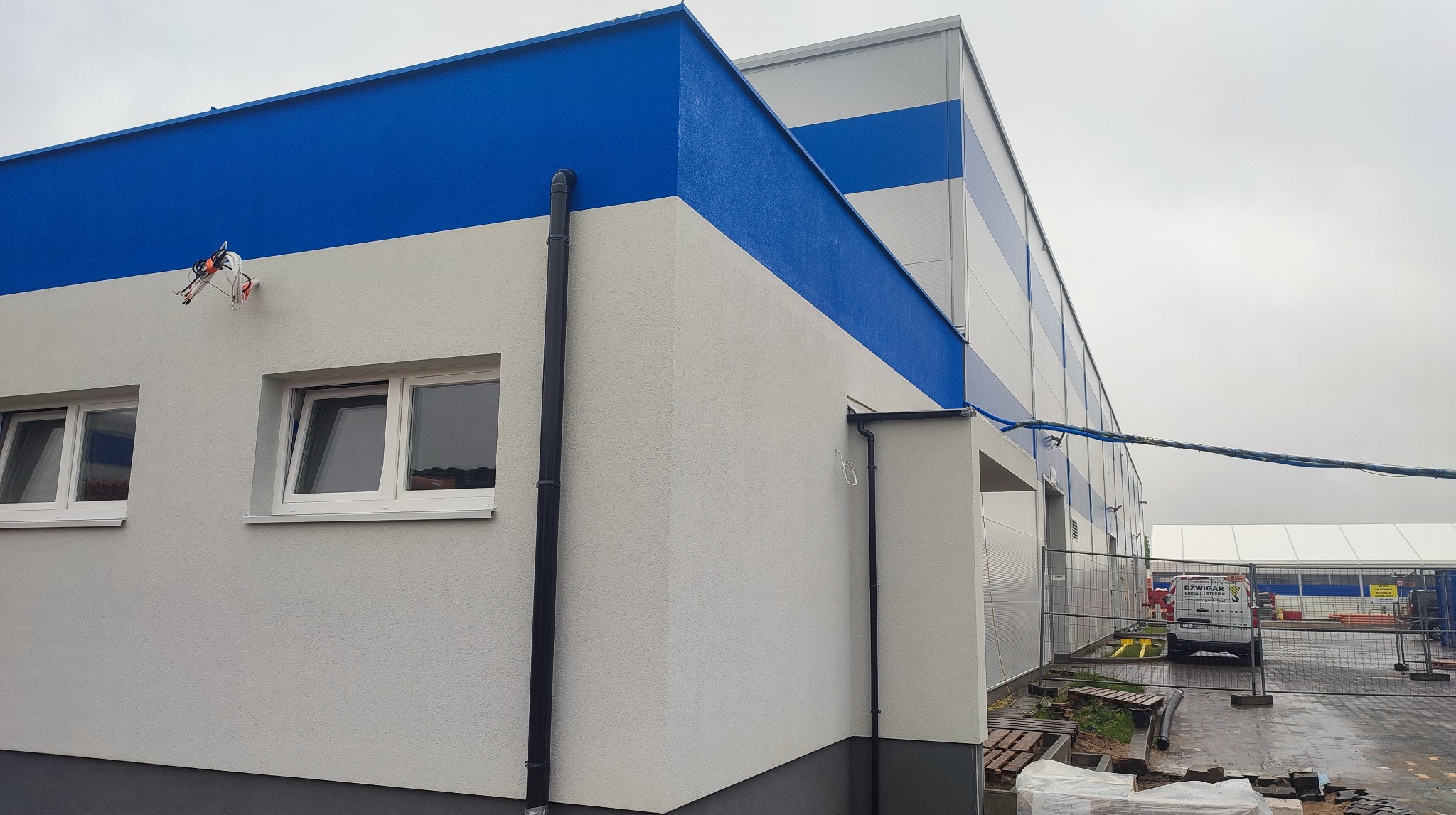 Wykończona elewacja budynku przemysłowego z wyraźnym podziałem na białą i niebieską strefę kolorystyczną. Widoczne okna, rynny i fragment zaplecza budowy.