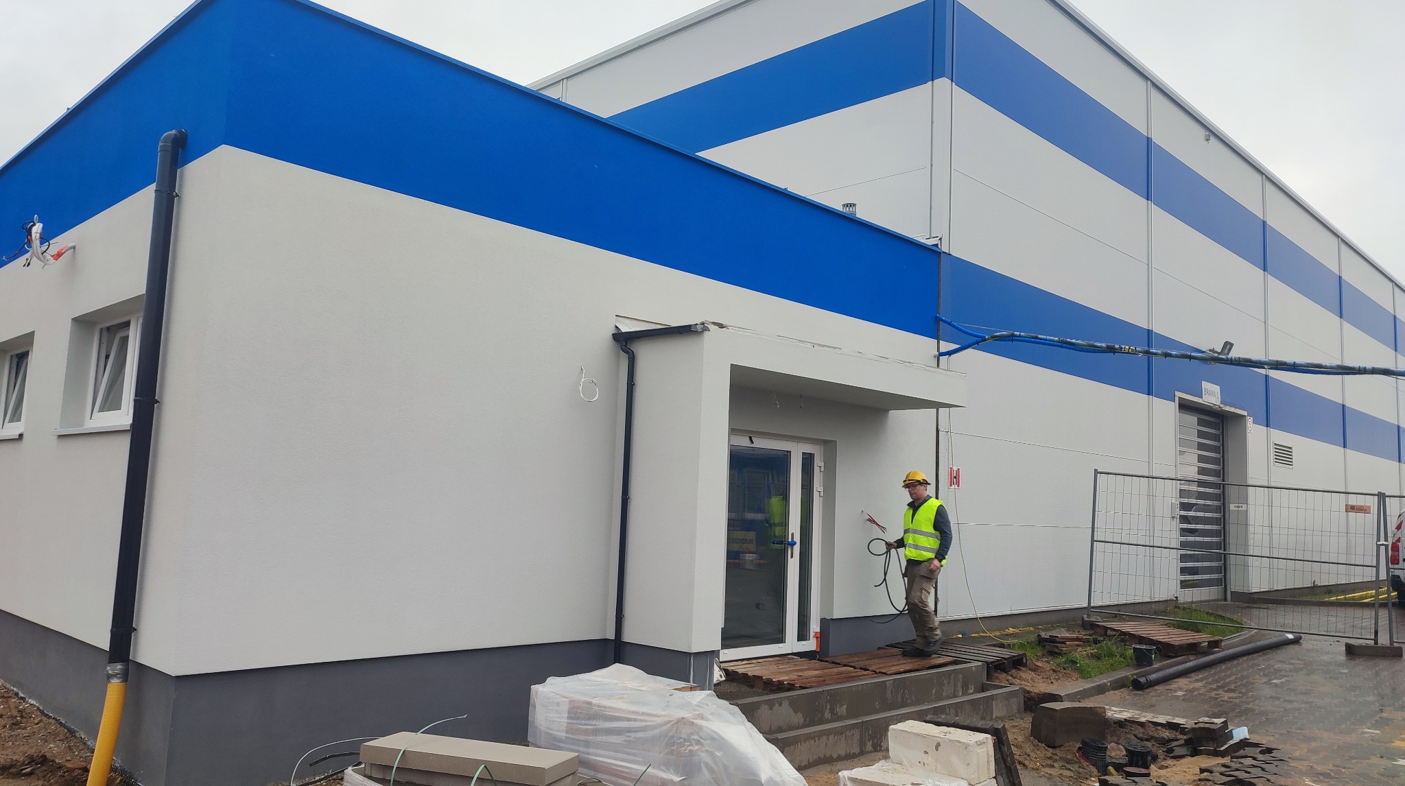 Pracownik w żółtym kasku, z pistoletem natryskowym, maluje elewację budynku w biało-niebieskie pasy. Widoczne okna, drzwi i elementy konstrukcyjne.