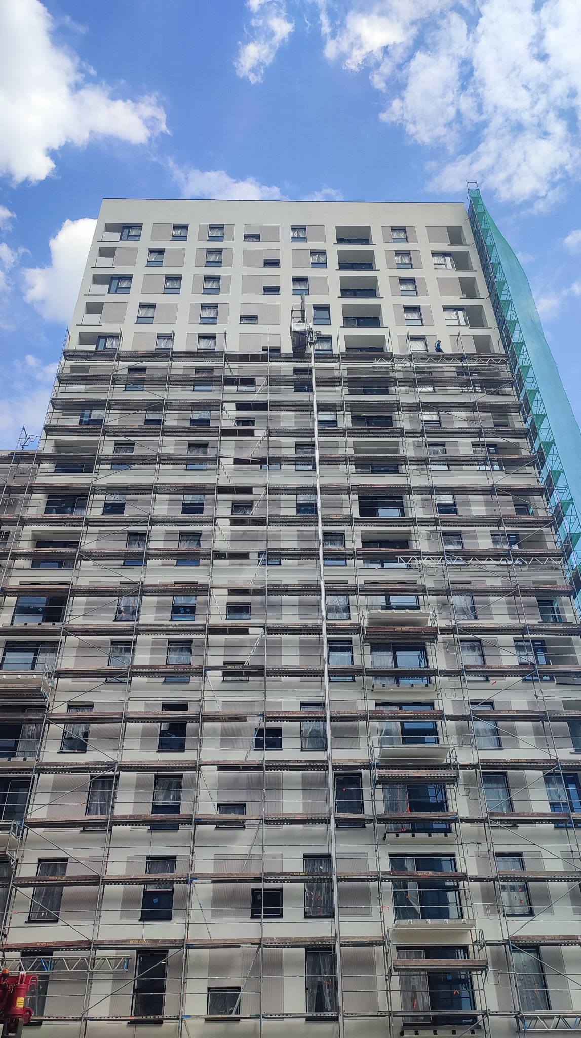 Wysoki budynek mieszkalny w trakcie prac elewacyjnych, osłonięty rusztowaniami. Widoczne fragmenty siatki ochronnej i pracownik na górnych piętrach.