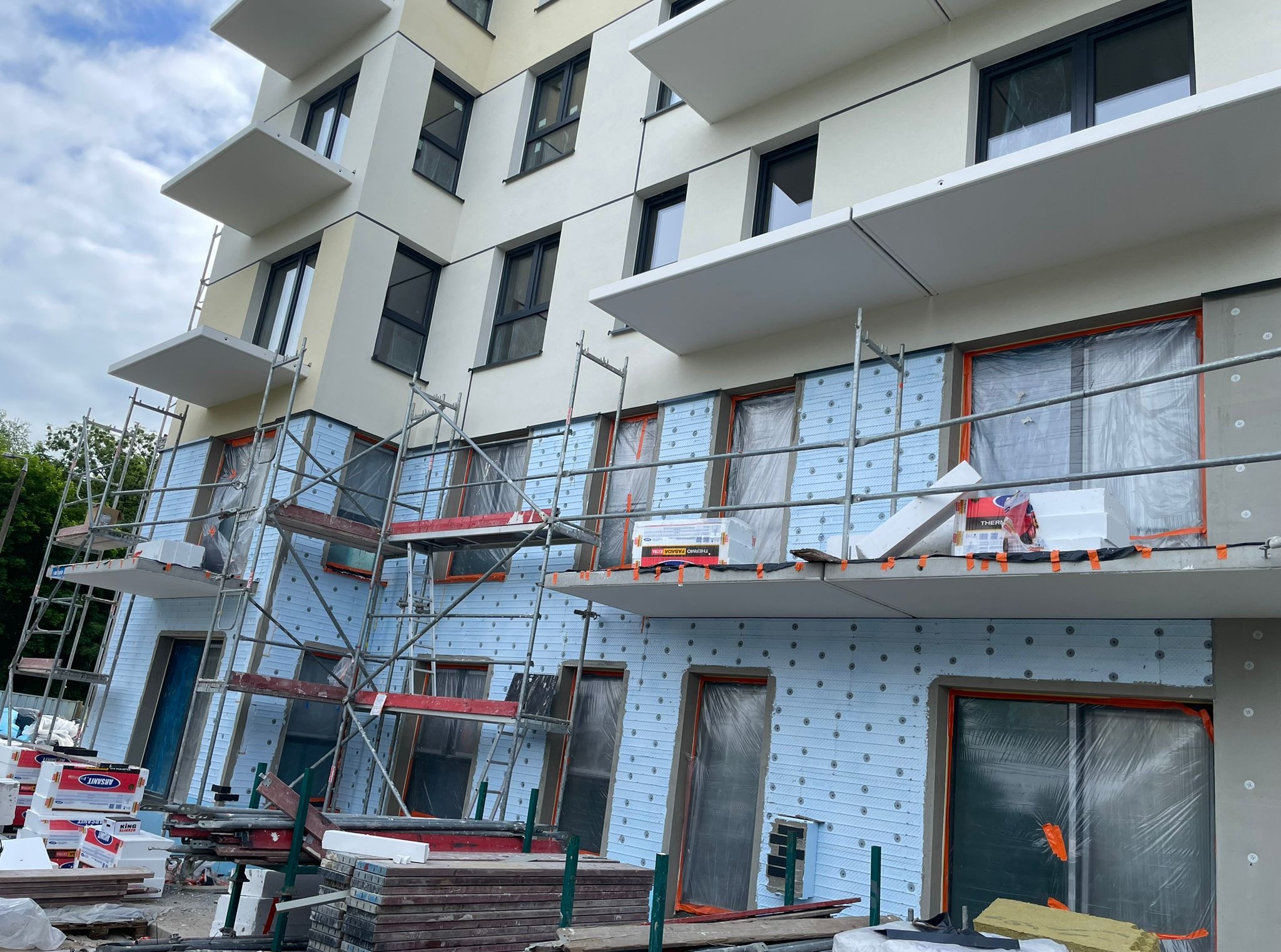 Ocieplanie elewacji budynku wielorodzinnego z balkonami, widoczne rusztowania i materiały izolacyjne. Okna zabezpieczone folią.