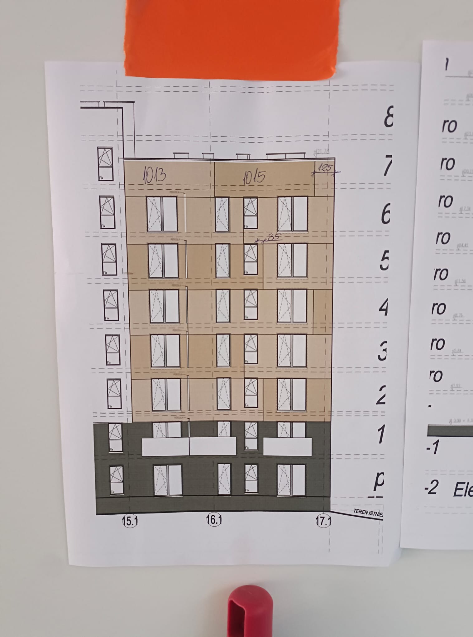 Plan elewacji budynku wielorodzinnego z numerami i wymiarami, przyczepiony do ściany pomarańczową taśmą. Widoczne detale architektoniczne i oznaczenia techniczne.