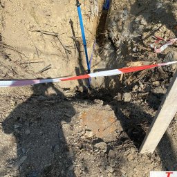 InstalDesign Engineering Paweł Liberda - Wykop z niebieskimi rurami instalacji sanitarnych, zabezpieczony taśmą ostrzegawczą. Widoczny cień osoby i fragment deski. Prace ziemne w toku.