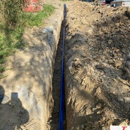 InstalDesign Engineering Paweł Liberda - Wykop z ułożonymi niebieskimi rurami instalacji sanitarnej na tle budowy domu jednorodzinnego. Widoczne materiały budowlane i sprzęt.