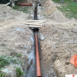 InstalDesign Engineering Paweł Liberda - Pomarańczowa rura kanalizacyjna w wykopie, widok z góry. Na ziemi deski, w tle zielona trawa i drzewa. Etap prac instalacyjnych.