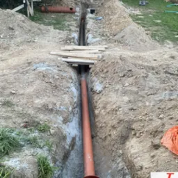 Pomarańczowa rura kanalizacyjna w wykopie, widok z góry. Na ziemi deski, w tle zielona trawa i drzewa. Etap prac instalacyjnych.