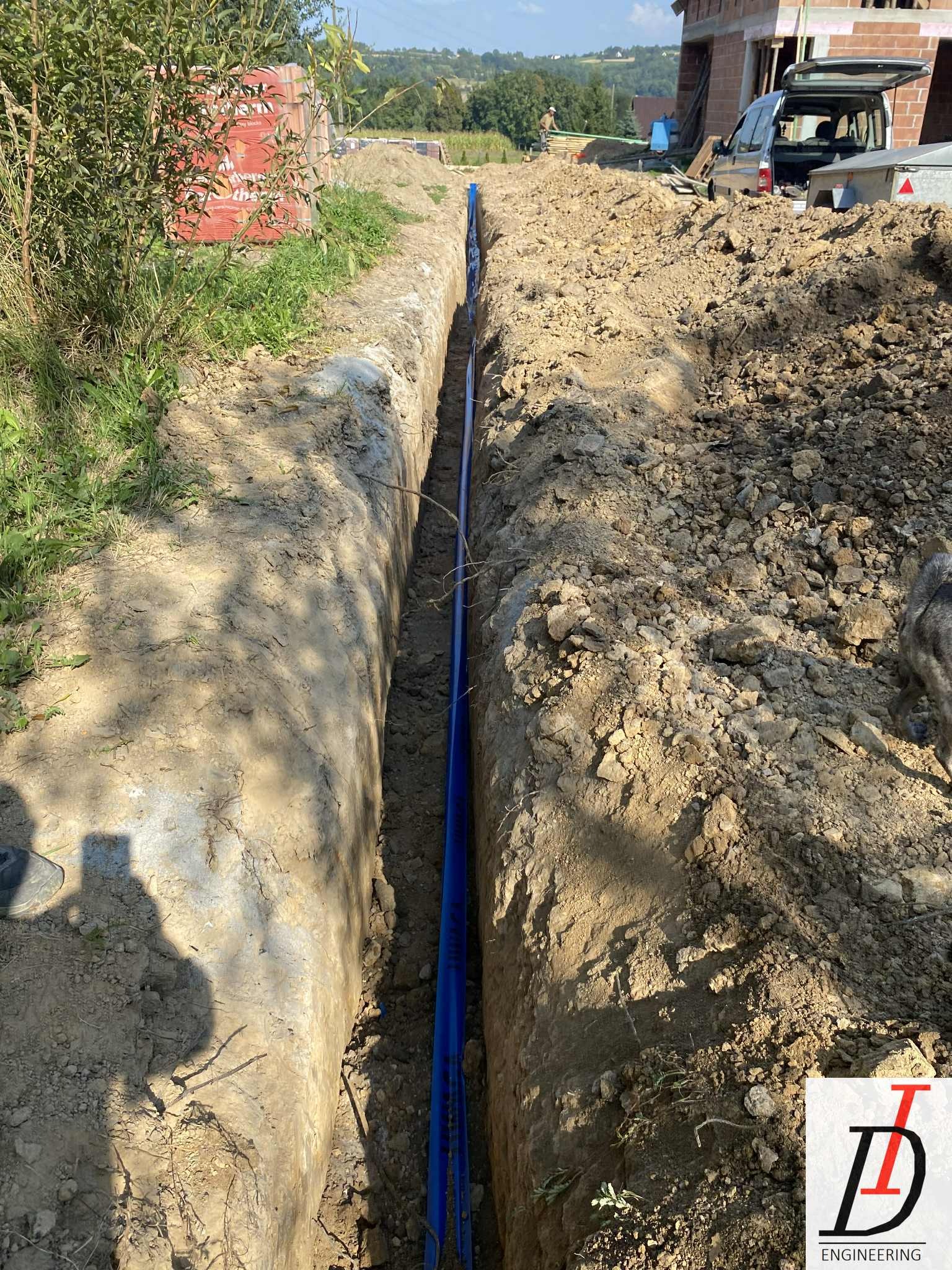 Wykop z ułożonymi niebieskimi rurami instalacji sanitarnej na tle budowy domu jednorodzinnego. Widoczne materiały budowlane i sprzęt.