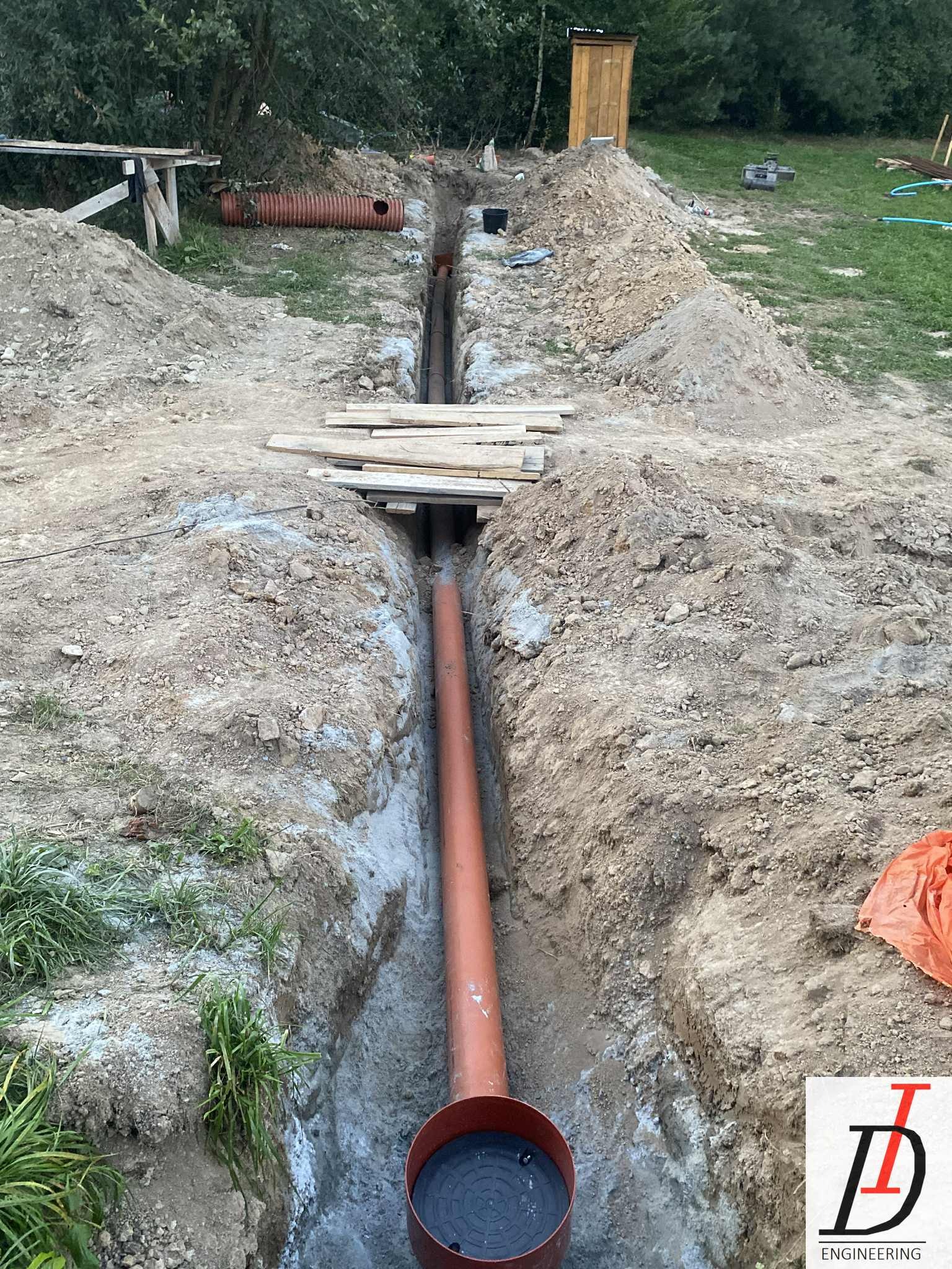 Pomarańczowa rura kanalizacyjna w wykopie, widok z góry. Na ziemi deski, w tle zielona trawa i drzewa. Etap prac instalacyjnych.