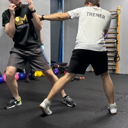 PM BOXING STUDIO SPÓŁKA Z OGRANICZONĄ ODPOWIEDZIALNOŚCIĄ - Trening boksu: mężczyzna w czarnej koszulce blokuje cios, trener w białej koszulce z napisem 'TRENER' wyprowadza uderzenie w sali z drabinkami i kettlebells.
