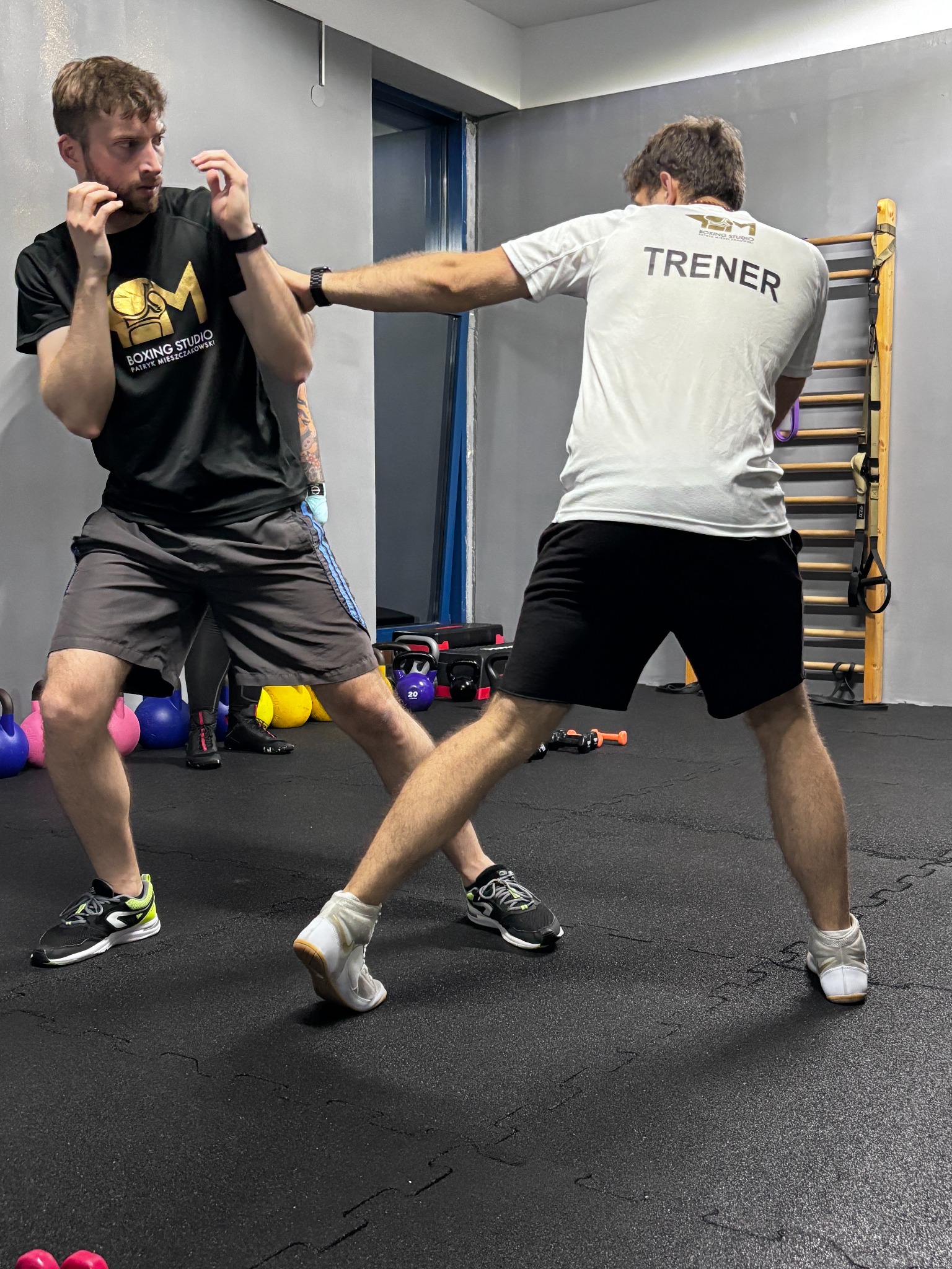 Trening boksu: mężczyzna w czarnej koszulce blokuje cios, trener w białej koszulce z napisem 'TRENER' wyprowadza uderzenie w sali z drabinkami i kettlebells.