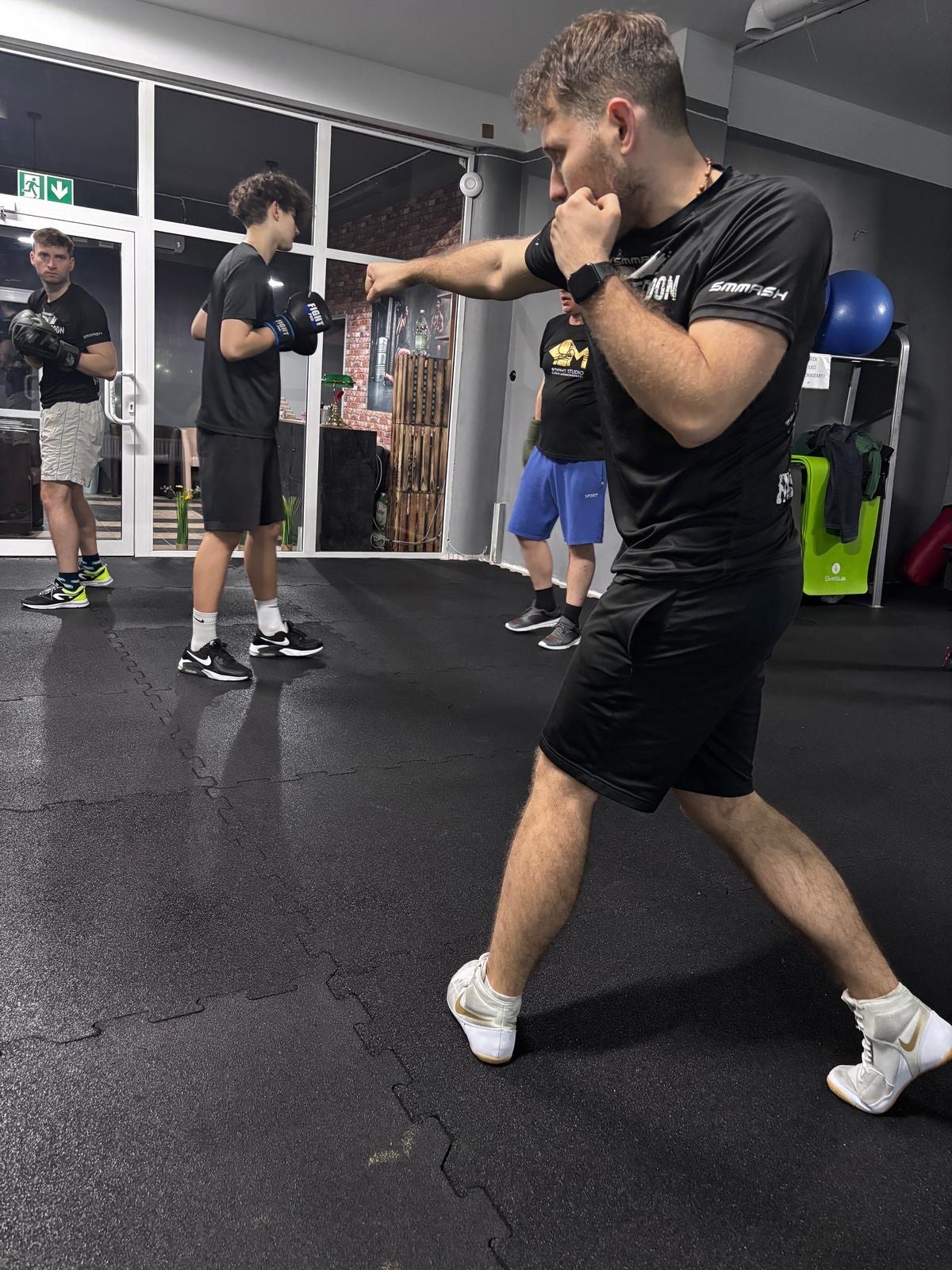 Mężczyzna w czarnym stroju bokserskim wyprowadza cios, w tle osoby w rękawicach i sprzęt sportowy na sali treningowej. Widoczne białe buty bokserskie.