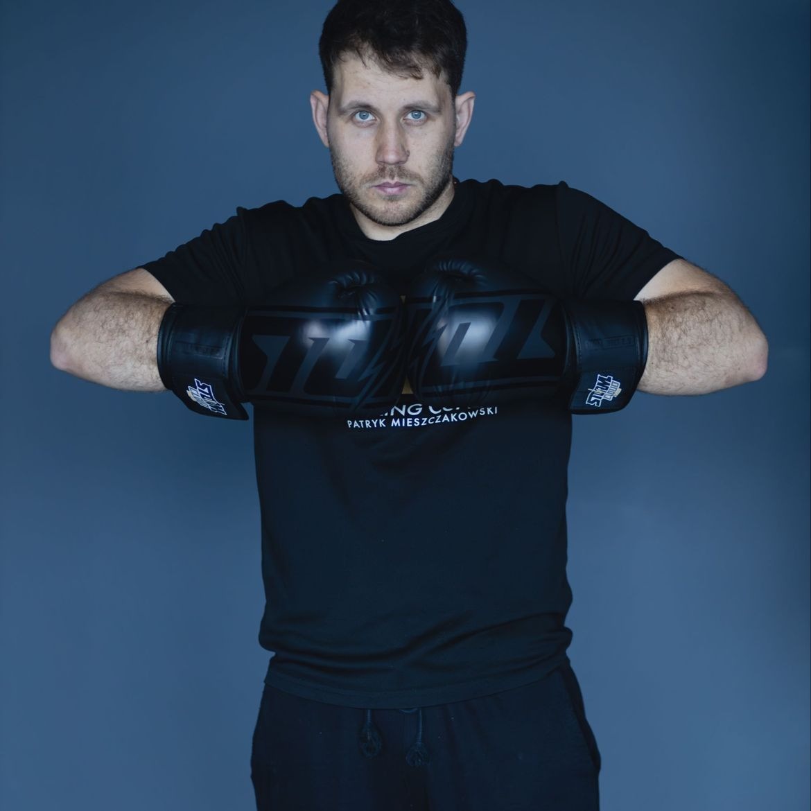 Mężczyzna z bokserkami na tle szarego studia, ubrany w czarny t-shirt z nadrukiem, prezentuje postawę gotową do walki, z powagą patrząc w obiektyw.