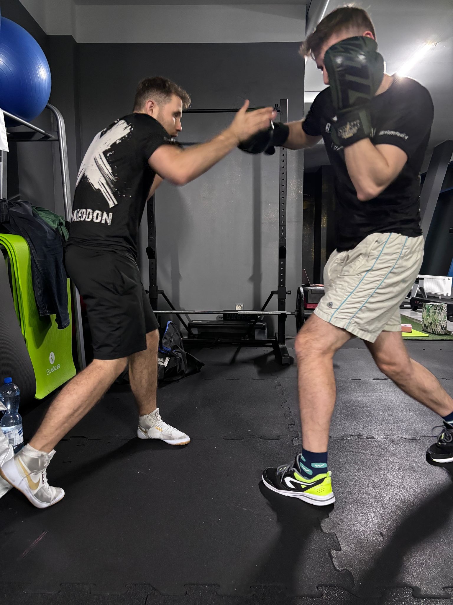 Mężczyźni w trakcie sparingu bokserskiego w siłowni. Jeden w czarnej koszulce z nadrukiem, drugi w rękawicach bokserskich.