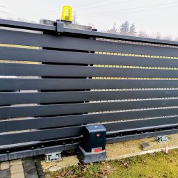 Paweł Luty Usługi Elektryczne - Nowoczesna, przesuwna brama w kolorze antracytu z żółtą lampą ostrzegawczą. Widoczny napęd bramy i elementy montażowe. Tło: zieleń trawy i drzewa.