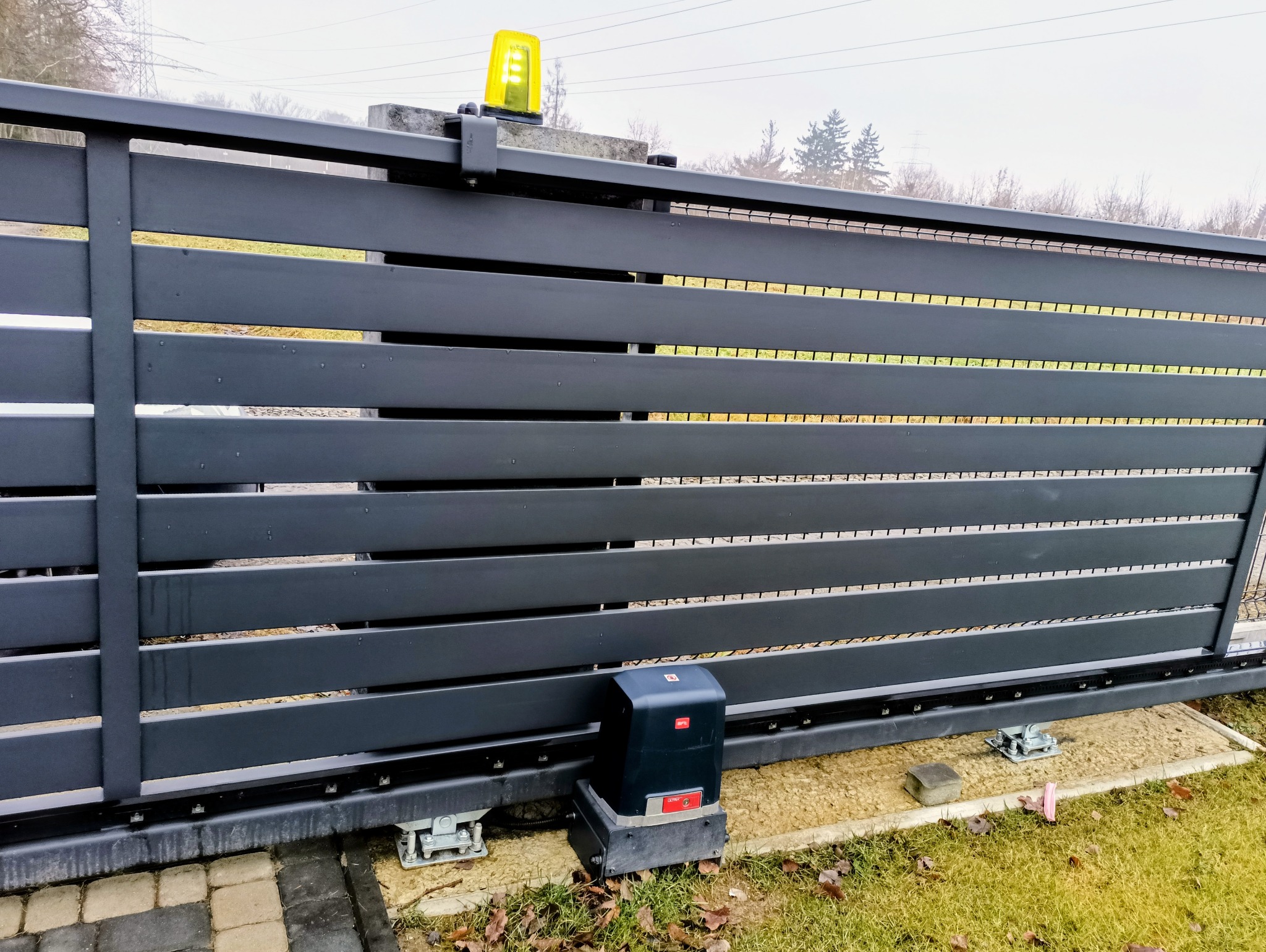 Nowoczesna, przesuwna brama w kolorze antracytu z żółtą lampą ostrzegawczą. Widoczny napęd bramy i elementy montażowe. Tło: zieleń trawy i drzewa.