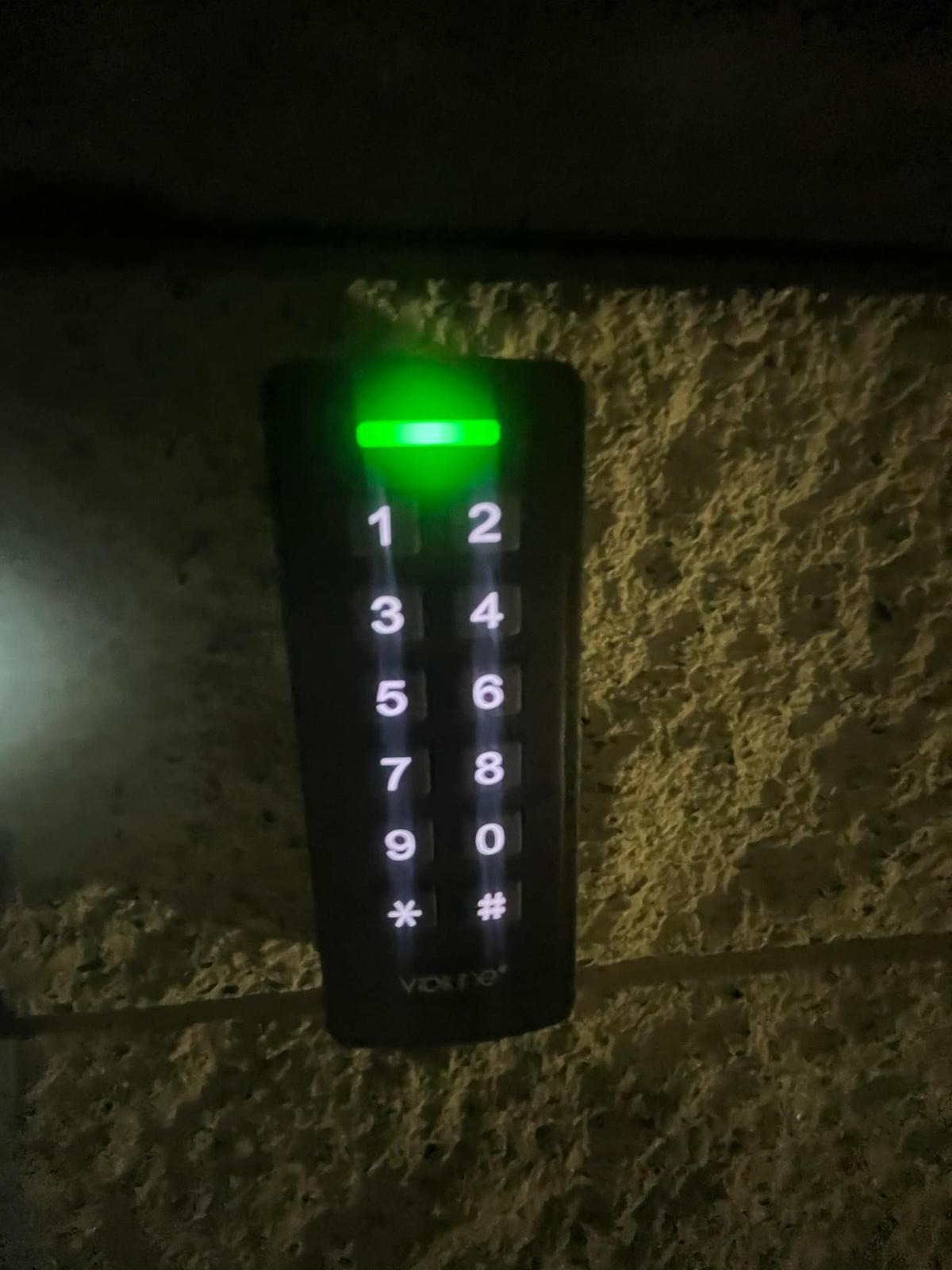 Zewnętrzny panel numeryczny domofonu z podświetlanymi cyframi i zieloną diodą LED, zamontowany na szorstkiej, jasnej ścianie. Widoczne oznaczenia cyfr i symboli.