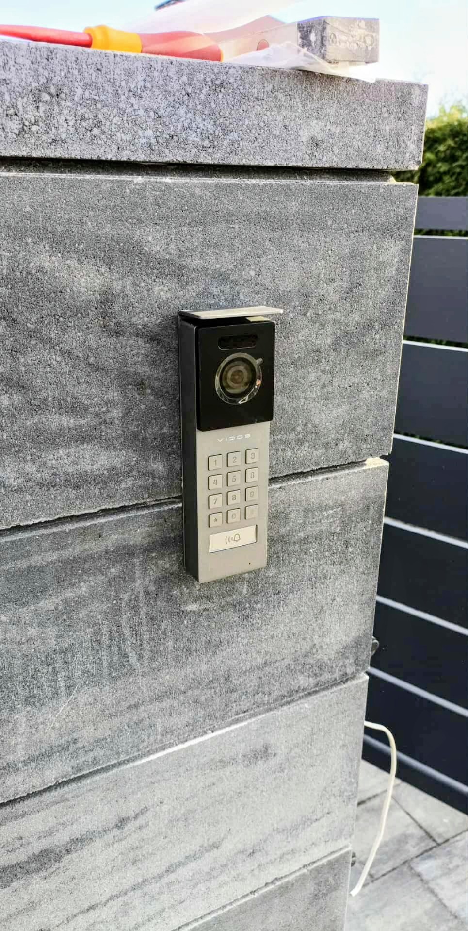 Szary wideodomofon z klawiaturą numeryczną, zamontowany na kamiennym słupku ogrodzenia. Widoczne narzędzia na górze słupka sugerują świeży montaż.