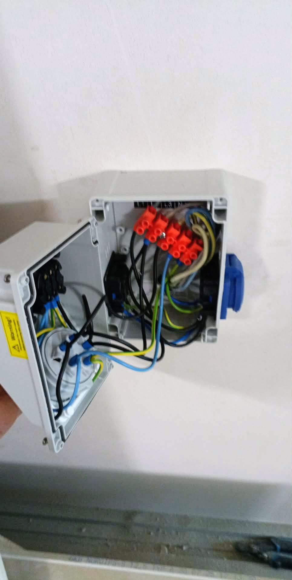 Otwarta puszka elektryczna z widocznymi przewodami, złączkami WAGO i elementami instalacji, zamontowana na białej ścianie. Widoczny fragment żółtej naklejki ostrzegawczej.