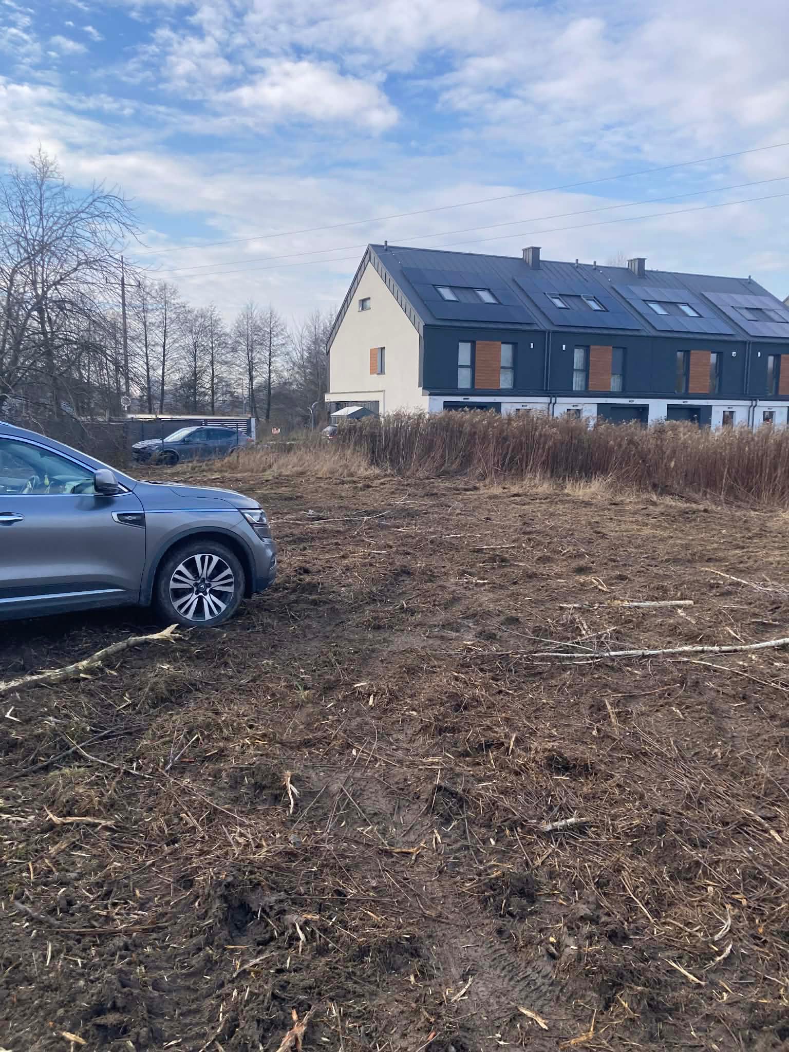 Teren po karczowaniu z widocznymi śladami opon, fragmentem auta i budynkami w tle. Poziom gruntu nierówny, z pozostałościami po roślinności.