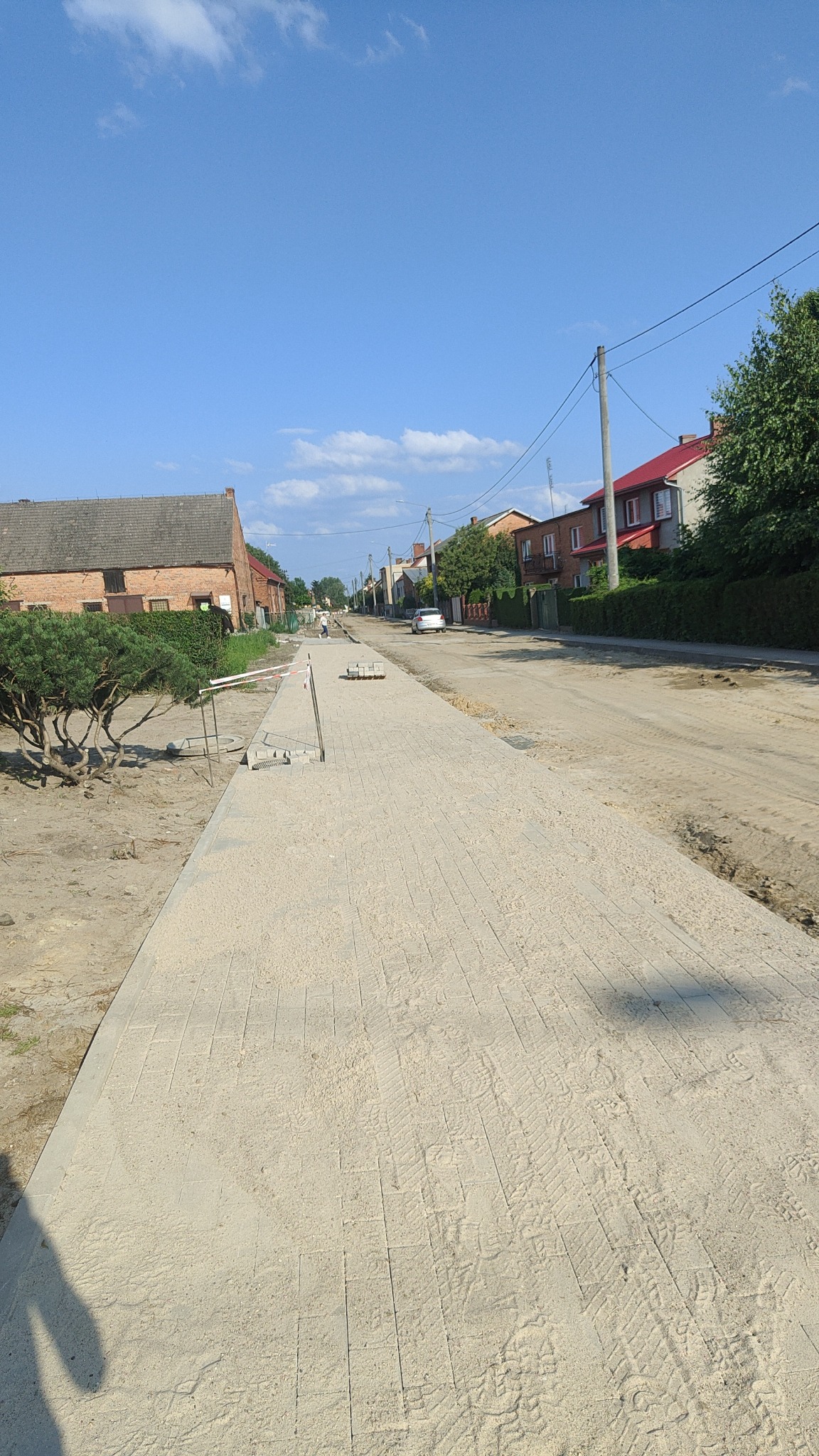 Ułożona kostka brukowa na ulicy w trakcie realizacji, widoczne przygotowane podłoże, resztki kostki i narzędzia. Perspektywa uliczna, zabudowa w tle.