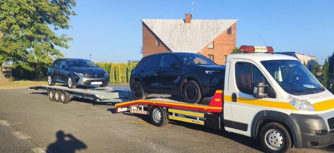 Laweta z dwoma samochodami osobowymi: szarym Renault i czarnym SUV-em Opel, na tle budynku z czerwonej cegły i zieleni. Pomoc drogowa.