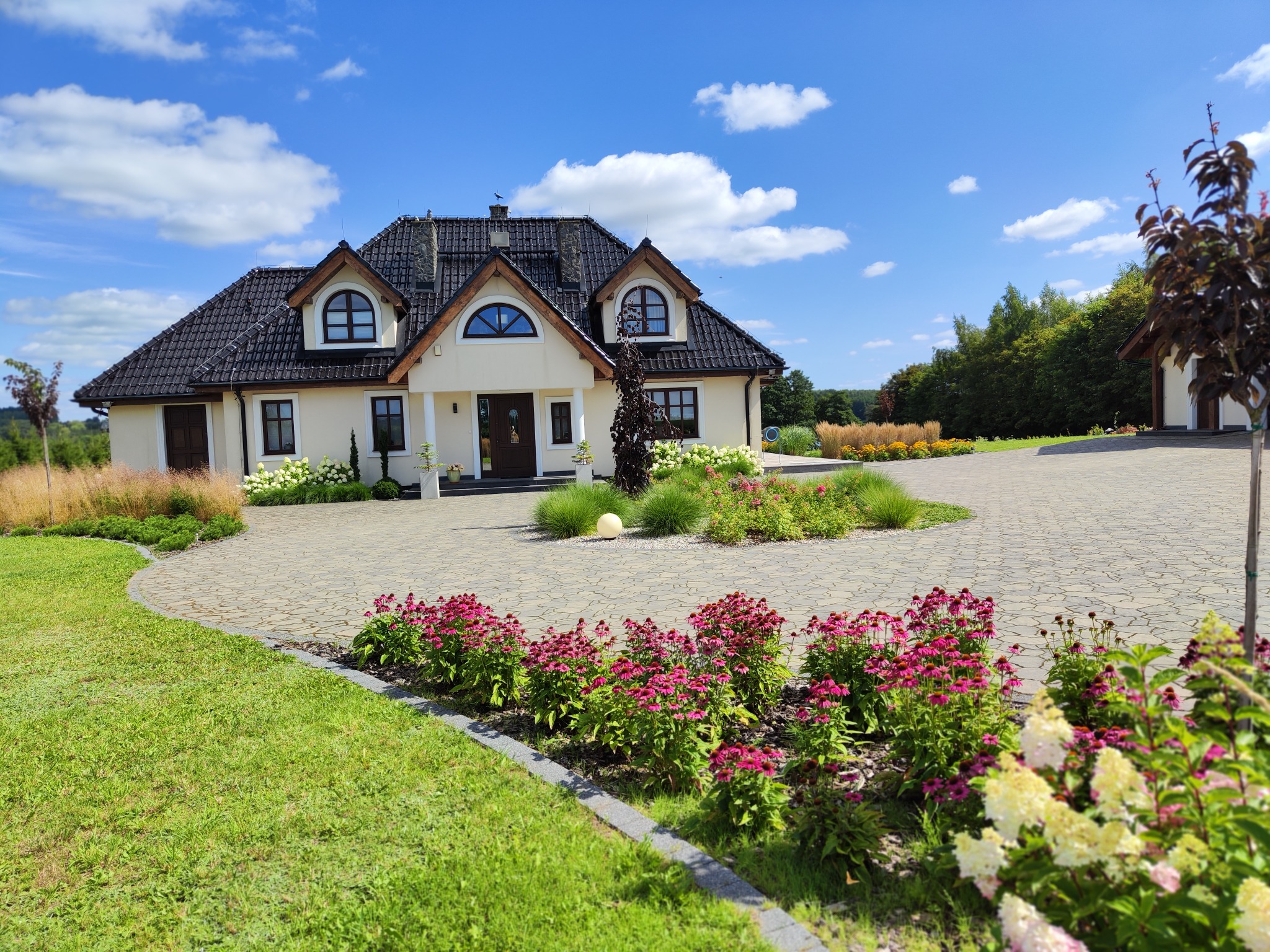 Ogród z podjazdem z kostki brukowej, kwiatami i trawnikiem przed domem z ciemnym dachem w słoneczny dzień. Widok na zadbany ogród i architekturę domu.