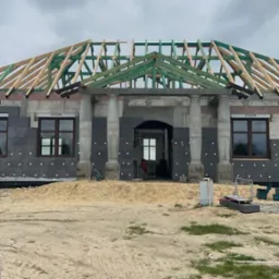Budowa domu w toku: widoczne ściany z izolacją, drewniana konstrukcja dachu, kolumny przy wejściu i okna z roletami. Etap surowy zamknięty.