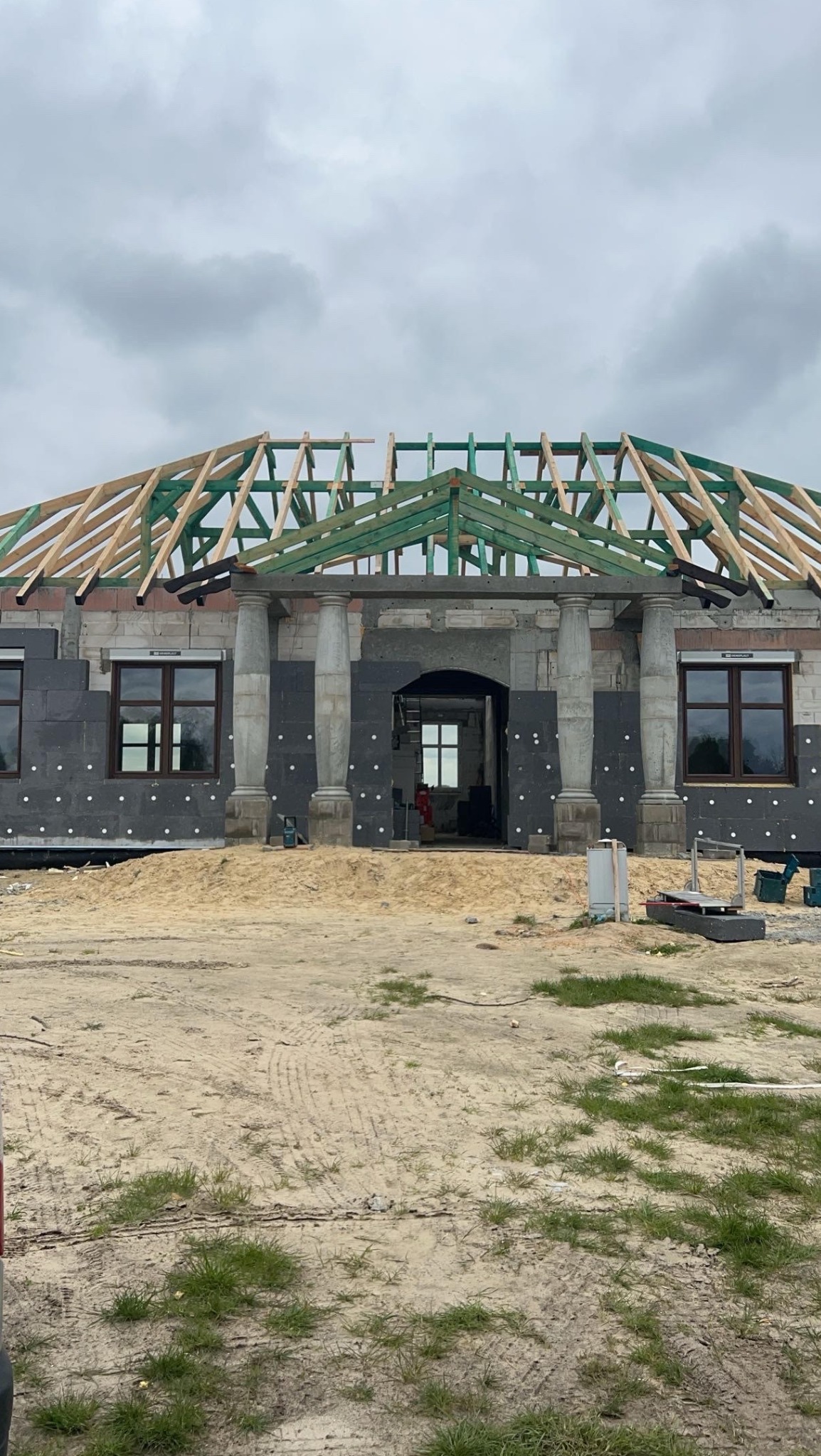 Budowa domu w toku: widoczne ściany z izolacją, drewniana konstrukcja dachu, kolumny przy wejściu i okna z roletami. Etap surowy zamknięty.