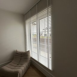 Eurofirany - Jasne, drewniane żaluzje poziome zamontowane w oknie z widokiem na dom. Obok okna beżowy, miękki fotel na drewnianej podłodze. Stonowana kolorystyka wnętrza.