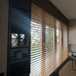 Eurofirany - Jasne, drewniane żaluzje poziome w łazience, z widokiem na zieleń za oknem. Minimalistyczny design, ciepłe oświetlenie.