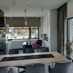 Eleganckie wnętrze: stół z szarymi krzesłami, w tle kuchnia z roletą w oknie i zasłoną przy oknie. Minimalistyczny design z akcentem roślinnym.