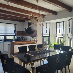 Eurofirany - Eleganckie rolety rzymskie w jasnym wnętrzu jadalni z czarnymi krzesłami i złotymi akcentami. Widoczne drewniane belki sufitowe i nowoczesna lampa.