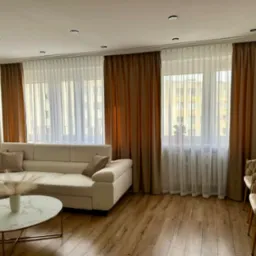 Salon z beżową sofą i marmurowym stolikiem. Okna przesłonięte brązowymi zasłonami i białymi firanami. Podłoga wyłożona panelami w odcieniach brązu.