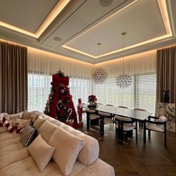 Eurofirany - Elegancki salon z beżowymi zasłonami i roletami wewnętrznymi, świąteczny wystrój z choinką i dekoracjami. Nowoczesne oświetlenie sufitowe.