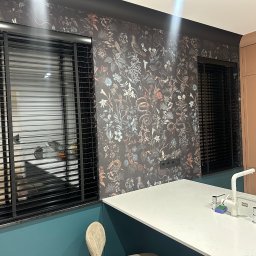 Eurofirany - Czarne żaluzje poziome w oknach kuchni z tapetą w kwiaty i wzory. Blat kuchenny z białego kamienia i bateria. Nowoczesne wykończenie wnętrza.