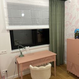 Eurofirany - Pokój dziecięcy z roletą rzymską, zieloną zasłoną i biurkiem w kolorze pudrowego różu. Na biurku lampka, a na szafce figurka dinozaura. Tapeta w motywy roślinne i zwierzęce.