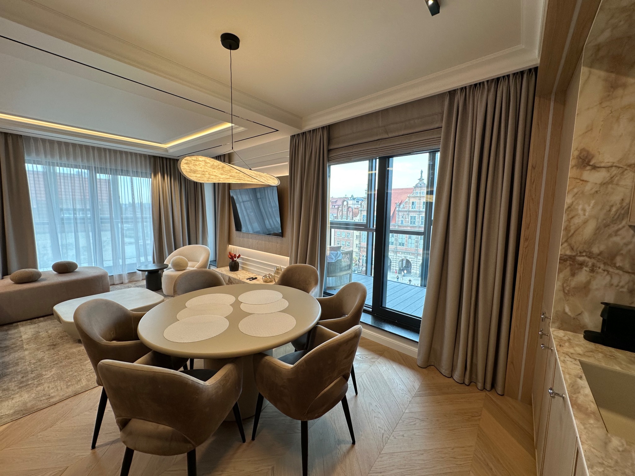 Eleganckie wnętrze: beżowe zasłony, roleta rzymska i widok na miasto. Stół z krzesłami w salonie, parkiet w jodełkę i designerska lampa.