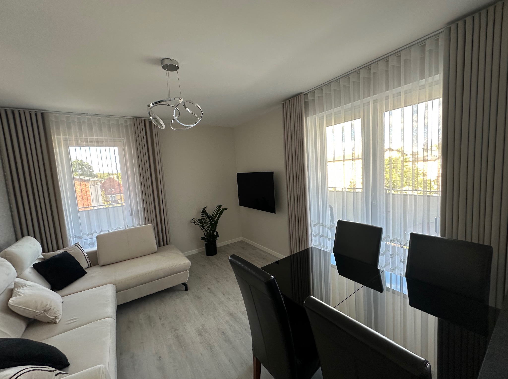 Jasne wnętrze salonu z beżowymi zasłonami i firanami w oknach. Widoczny narożnik, stół z krzesłami i nowoczesna lampa. Stonowana kolorystyka.